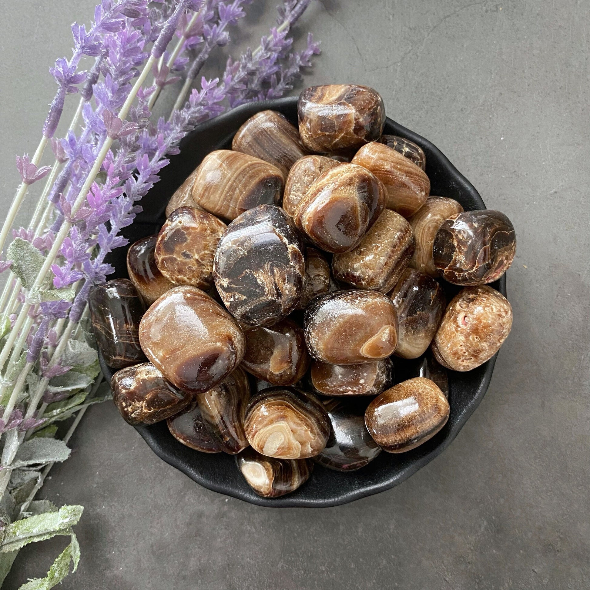 A black bowl filled with polished brown and beige stones sits on a gray surface. Beside the bowl, a sprig of purple lavender accentuates the natural tones of the gemstones. The glossy finish and varied patterns suggest they are Chocolate Calcite Tumbled Stones for grounding energy.