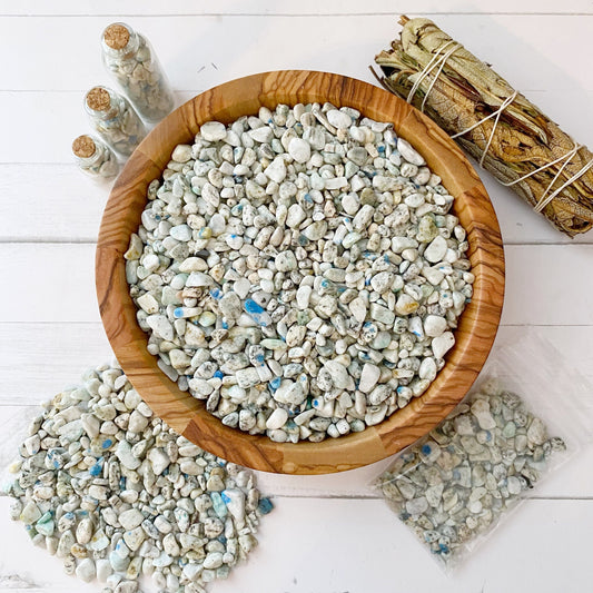 A wooden bowl filled with small, white stones with hints of blue and gray sits on a white surface. Nearby, there is a cluster of similar stones in a plastic bag labeled as K2 Jasper Crystal Chips, three small bottles filled with K2 Jasper Crystal Chips for crafting supplies or metaphysical uses, and a bundle of dried sage wrapped with twine.