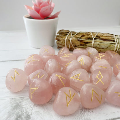 A collection of Rose Quartz Elder Futhark Runes is scattered on a white wooden surface. In the background, there is a small pink succulent in a white pot and a bundle of dried sage tied with twine.