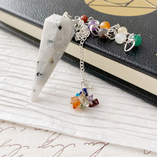 A Rainbow Moonstone Hexagonal Pendulum with a pointed, marbled white stone hangs over a textured surface. The connecting chain is adorned with colorful gemstone beads, perfect for balancing chakra crystals. The background features a black book with a gold geometric design on its cover.