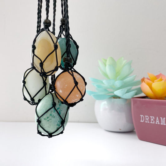 A collection of five polished stones of various colors, each encased in a handwoven black cord, hanging side by side. In the background, two small potted succulents with blue-green and orange leaves rest on a white surface. The product is called Empty Net Cage Necklace.