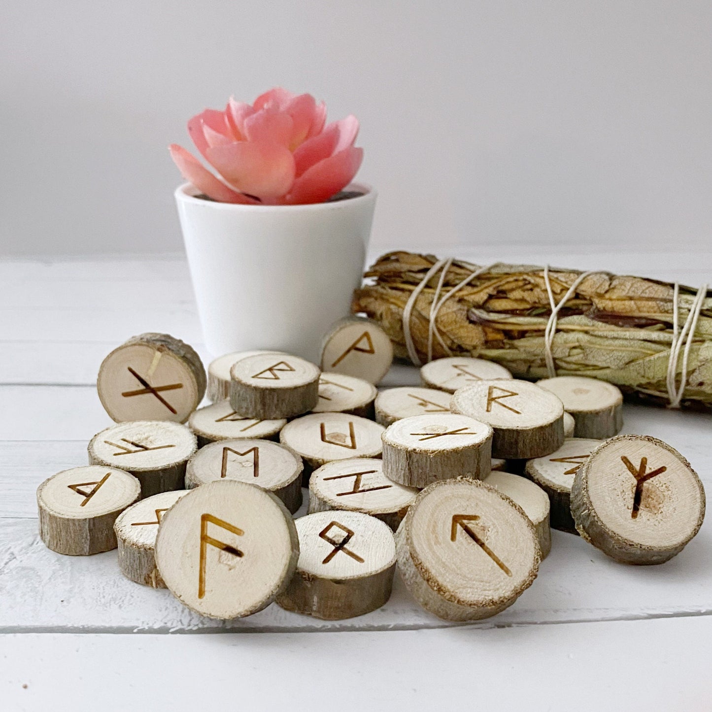 A collection of Poplar Wood Elder Futhark Runes with various symbols carved into them are scattered on a white surface. A small potted pink succulent is in the background, along with a bundle of dried sage, invoking the mystical traditions of Germanic tribes.