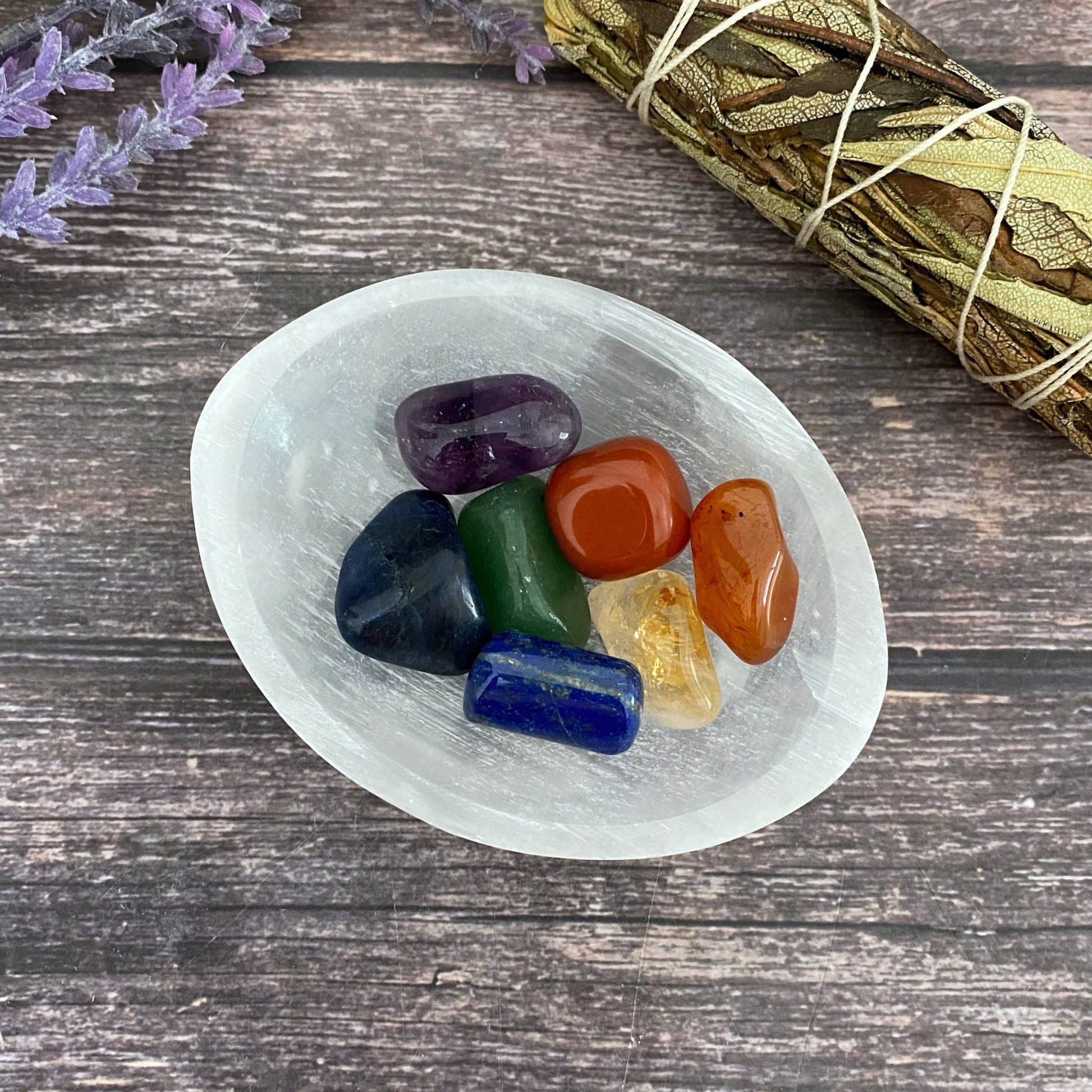 A white oval dish contains seven colorful polished stones, including amethyst, red jasper, citrine, and more. Next to the dish is a bundle of dried sage wrapped with string. The background consists of a wooden surface and sprigs of lavender. This setup serves as an elegant 4" Oval Selenite Charging Bowl.