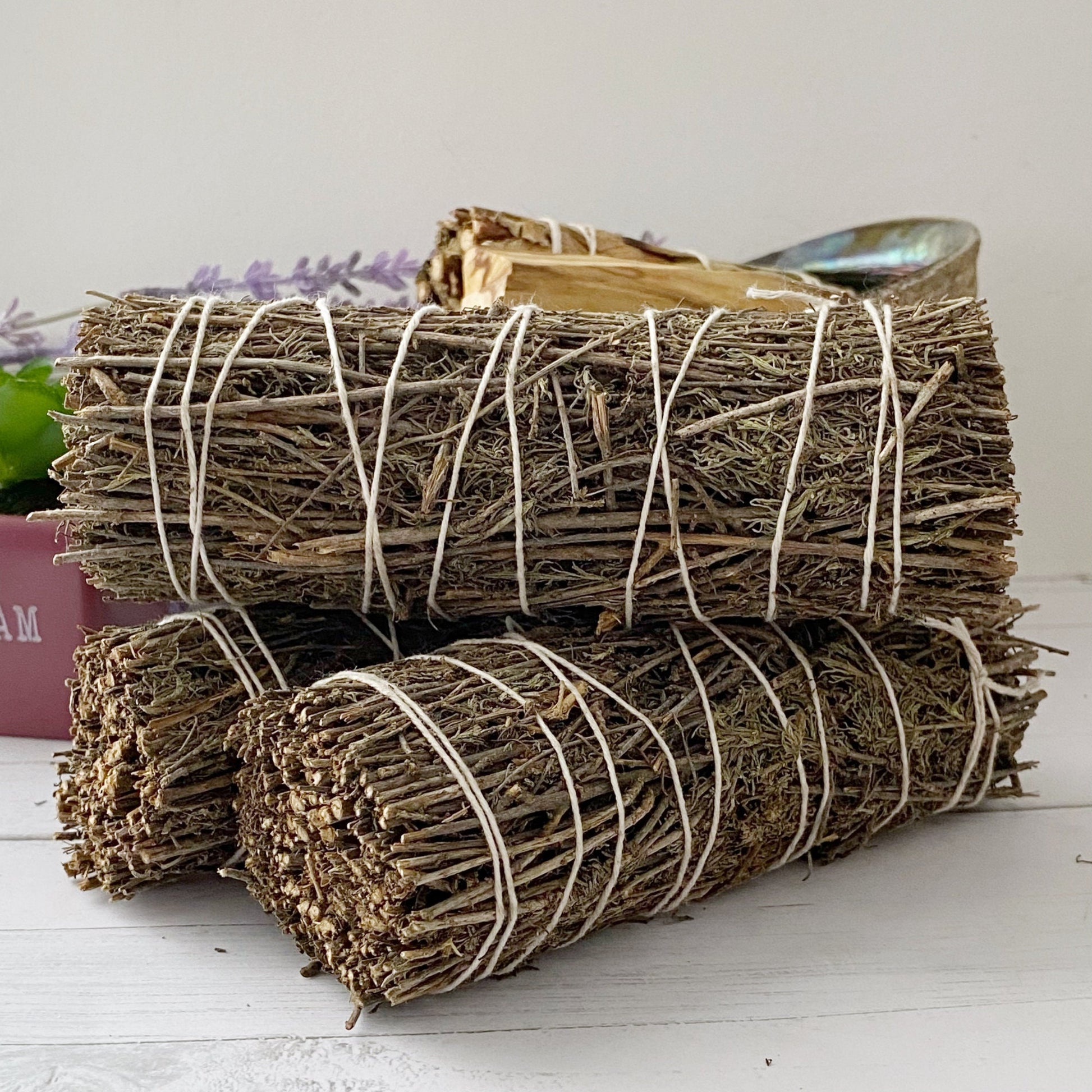 A close-up of three bundles of Organic Desert Sage Smudge Sticks tied with white string, placed on a white wooden surface. Behind them, a green succulent in a pink pot and a shell filled with desert sage and other dried herbs are visible. A lavender sprig adds a touch of color in the background.