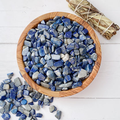 A wooden bowl filled with polished blue, navy, and white stones, including Lapis Lazuli Crystal Chips, is placed on a white wooden surface. Some stones are scattered outside the bowl. A bundle of sticks tied with string lies next to the bowl, perfect for metaphysical crafting or enhancing the Third Eye Chakra.