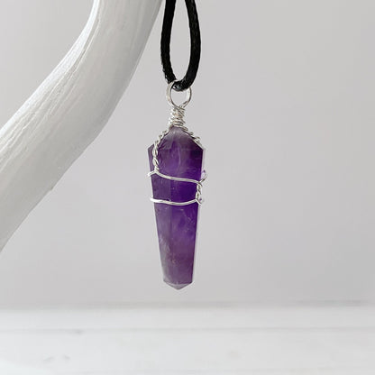 A close-up image of a 1.5" Double Terminated Amethyst Point Pendant featuring a polished, double-terminated stone wrapped with thin silver wire. Known for promoting emotional balance, the pendant is suspended from a black cord and displayed against a plain gray background.