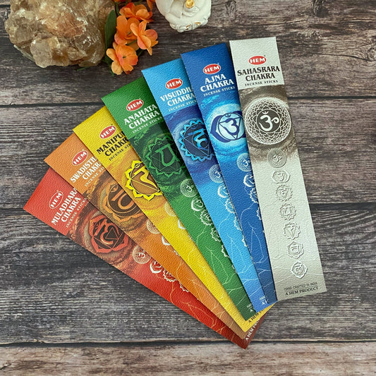 A 7 Chakra Incense Set arranged in a fan shape on a wooden surface. Each pack is colored differently, representing the chakras for meditation and Chakra balancing: Muladhara (red), Svadhisthana (orange), Manipura (yellow), Anahata (green), Vishuddha (blue), Ajna (indigo), and Sahasrara (purple).