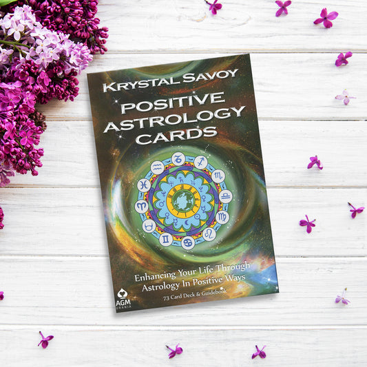 A deck of Positive Astrology Cards rests on a white wooden surface surrounded by clusters of small purple flowers and sprigs of lilac. The deck promises to enhance life through astrology, featuring insights into Zodiac Sun Signs, and includes 73 cards and a guidebook.