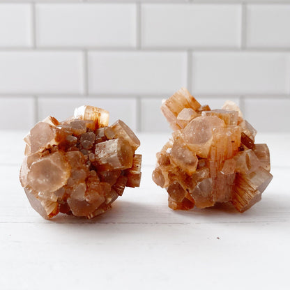 Two clusters of Raw Aragonite Cluster rest on a white surface with white subway tile in the background. The hexagonal shapes appear translucent, showcasing their intricate geometric formations, perfect for chakra balancing and grounding stone practices.