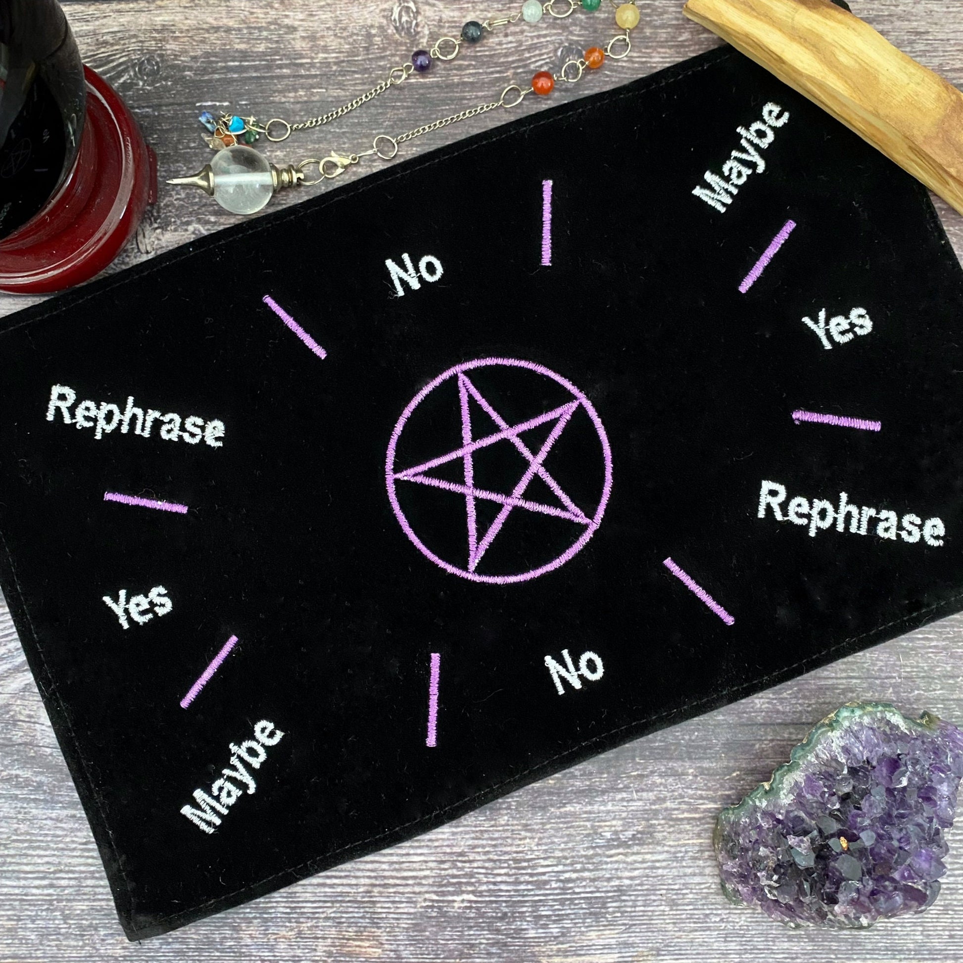 A Velvet Pentagram Pendulum Mat rests on a table with a gemstone necklace and amethyst cluster nearby. The black pendulum mat, adorned with a purple pentagram at the center, has "Yes," "No," "Maybe," and "Rephrase" written in white around it, each word highlighted by small rays.