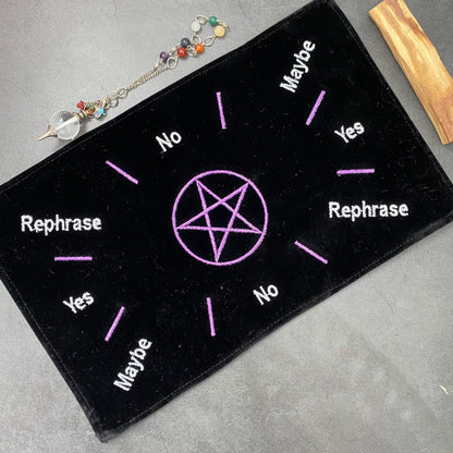 A Velvet Pentagram Pendulum Mat rests on a table with a gemstone necklace and amethyst cluster nearby. The black pendulum mat, adorned with a purple pentagram at the center, has "Yes," "No," "Maybe," and "Rephrase" written in white around it, each word highlighted by small rays.