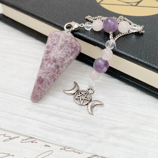 A Lepidolite Crystal Hexagonal Pendulum with Triple Moon Charm is placed on a black book with gold geometric designs. A Triple Moons charm featuring a pentacle is attached to the divination tool. The background includes handwritten notes.