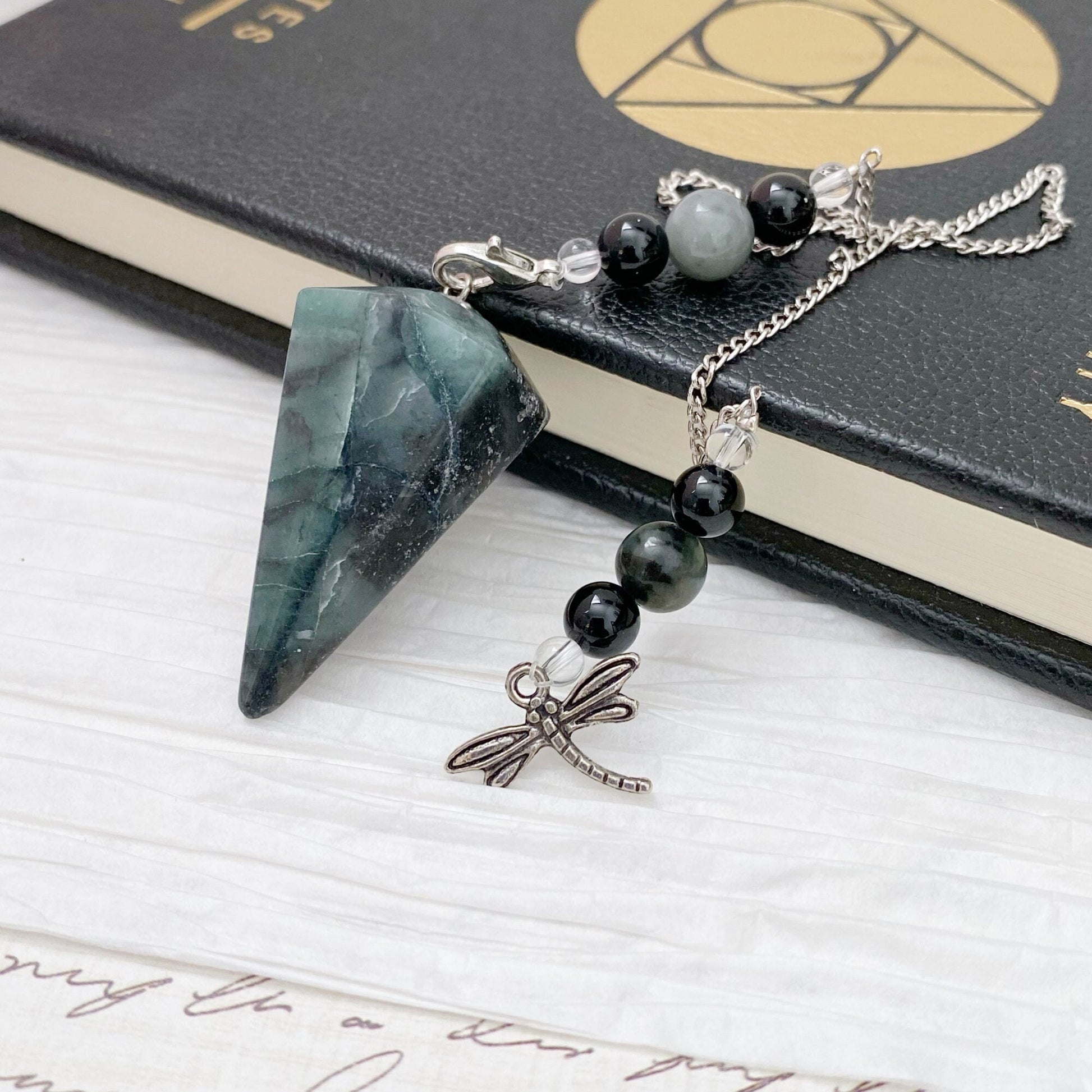 An Emerald Hexagonal Pendulum with Dragonfly Charm is placed on an open book with a black cover. The chain of the pendulum is adorned with various black onyx beads. The setup rests on a white textured surface with slight rustic elements visible.