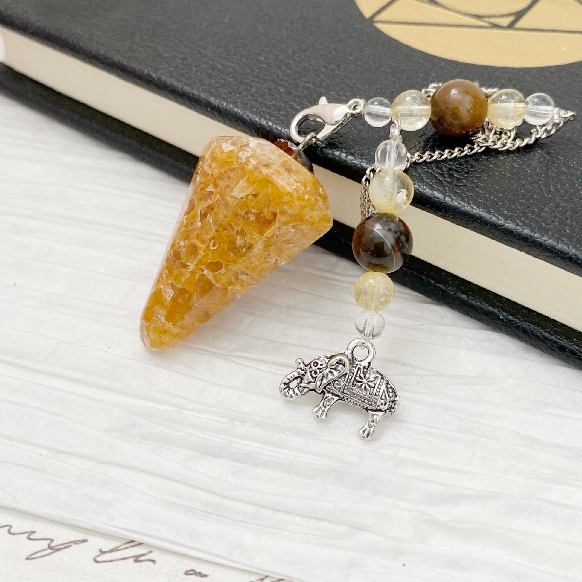 A Golden Healer Quartz Hexagonal Pendulum with Lucky Elephant, featuring a yellow crystal and multicolored beads, is laid on a black notebook. The chain includes a small, ornate silver elephant charm, adding a decorative touch. Ideal for divination or dowsing, the Golden Healer Quartz Hexagonal Pendulum with Lucky Elephant promotes crystal healing.