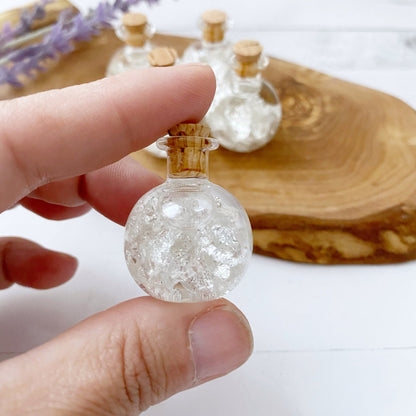 A .999 Silver Flakes Bottle filled with crystal-like white flakes and topped with a cork stopper, arranged on a wooden surface. In the background, there's a soft-focus sprig of purple lavender flowers.