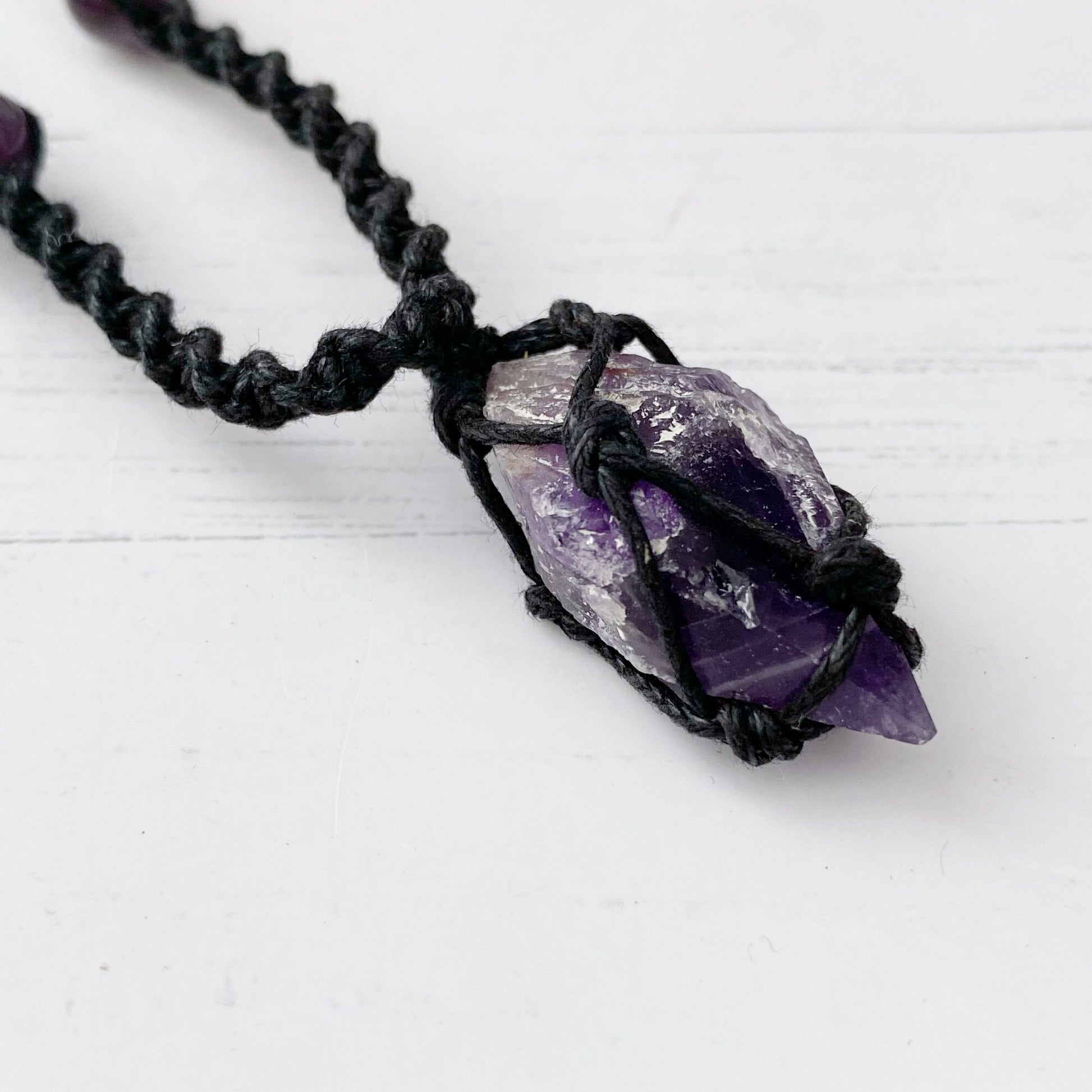 An Amethyst Crystal Point Net Cord Necklace displayed on a beige bust. The necklace features purple and white beads evenly spaced along the cord. The centerpiece is a larger, irregularly shaped purple stone wrapped in black macrame, enhancing spiritual awareness.