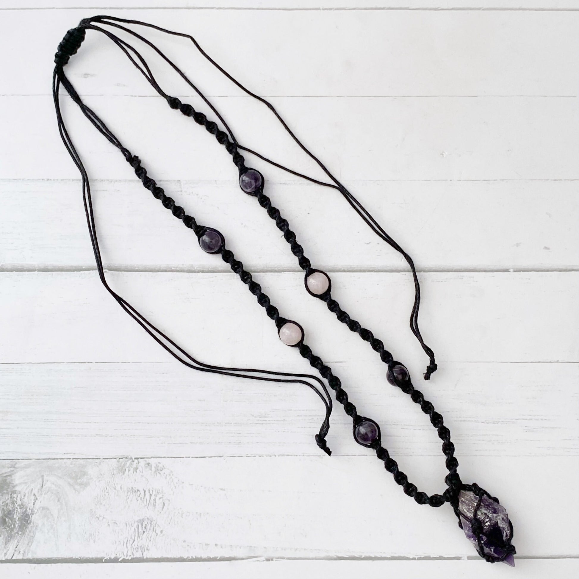 An Amethyst Crystal Point Net Cord Necklace displayed on a beige bust. The necklace features purple and white beads evenly spaced along the cord. The centerpiece is a larger, irregularly shaped purple stone wrapped in black macrame, enhancing spiritual awareness.