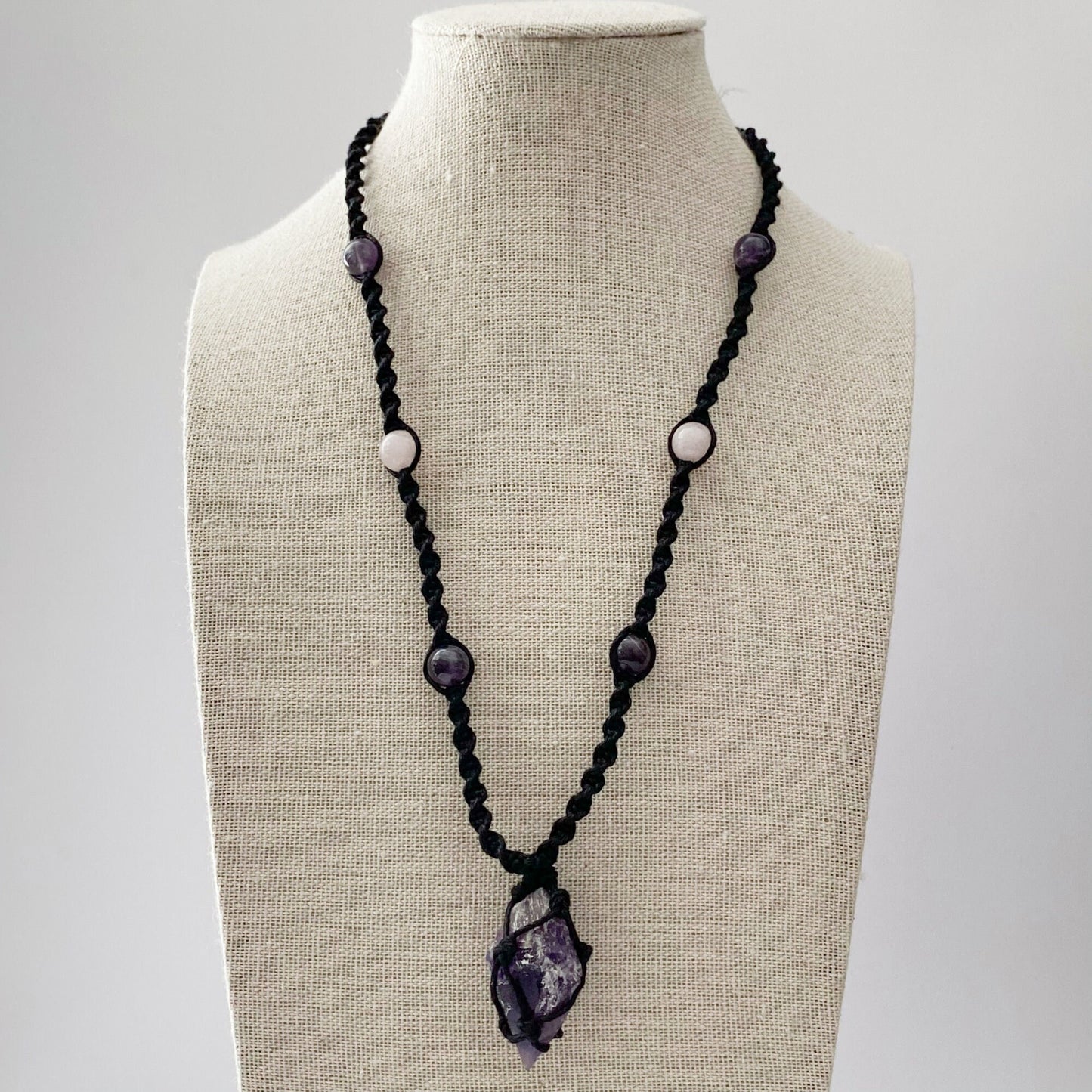 An Amethyst Crystal Point Net Cord Necklace displayed on a beige bust. The necklace features purple and white beads evenly spaced along the cord. The centerpiece is a larger, irregularly shaped purple stone wrapped in black macrame, enhancing spiritual awareness.