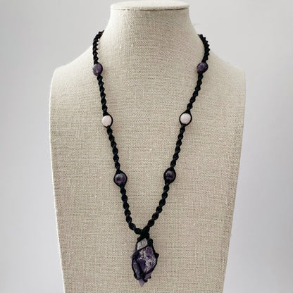 An Amethyst Crystal Point Net Cord Necklace displayed on a beige bust. The necklace features purple and white beads evenly spaced along the cord. The centerpiece is a larger, irregularly shaped purple stone wrapped in black macrame, enhancing spiritual awareness.