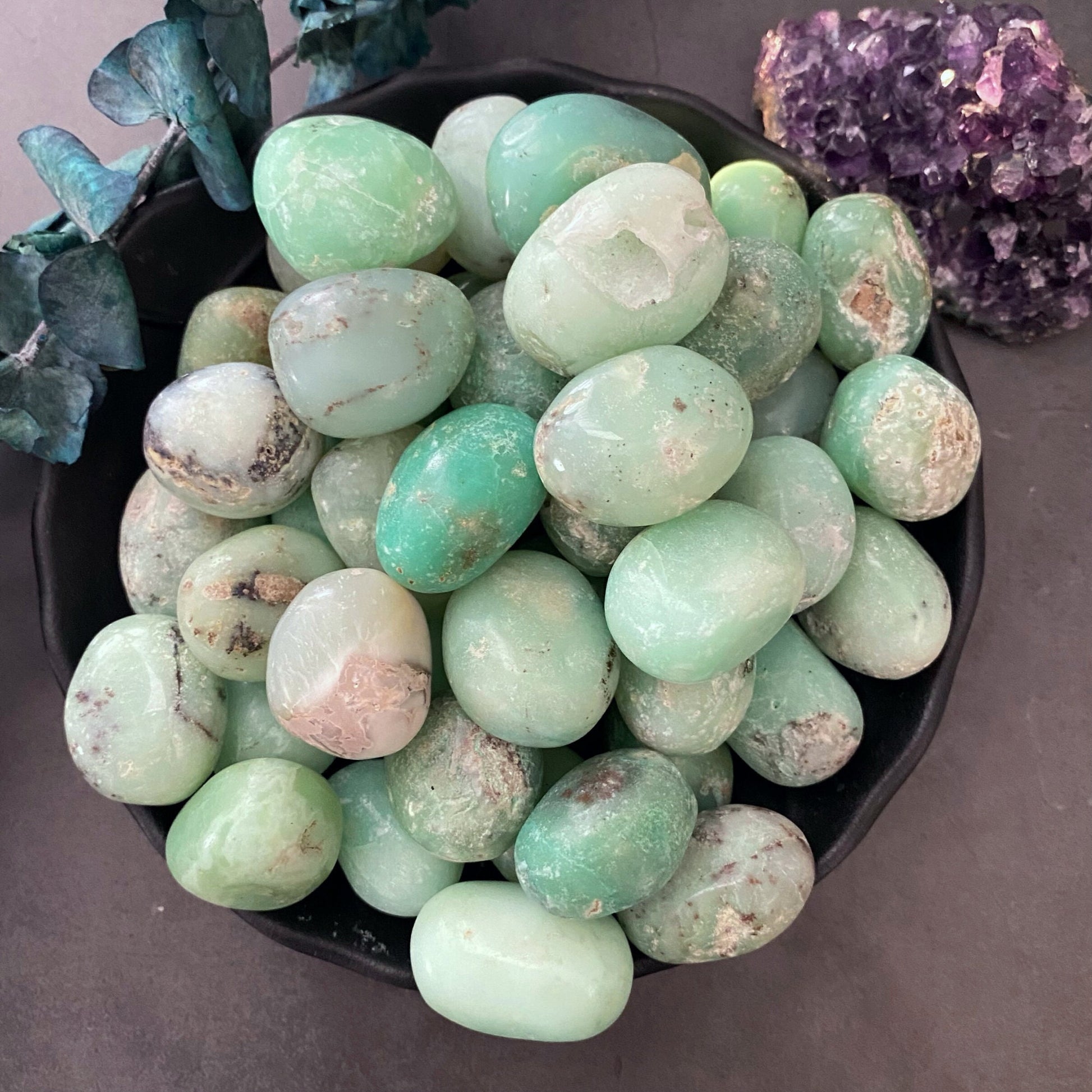 A black bowl filled with polished, oval green Chrysoprase Tumbled Stones varying in shades and patterns sits on a dark surface. To the left, a sprig of red berries and green leaves adds a touch of color and natural contrast, promoting creativity and emotional healing.