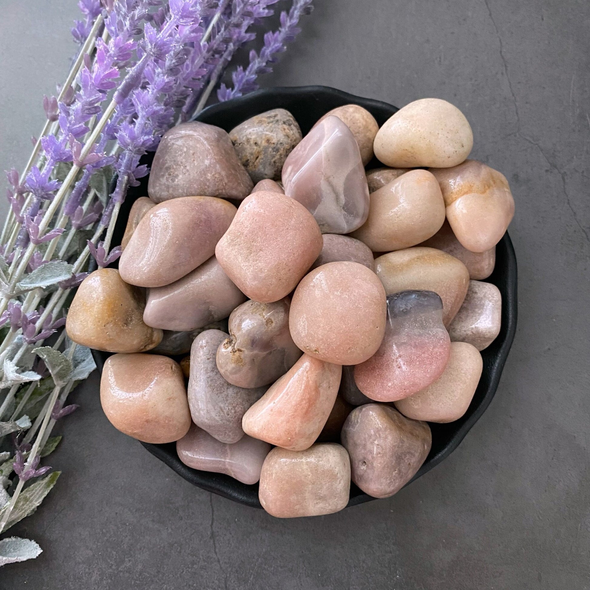 A black bowl filled with smooth, polished Pink Amethyst Tumbled Stones sits on a dark gray surface. To the left of the bowl, a sprig of dried lavender adds a touch of purple color and texture. The stones and lavender create a calming and soothing composition.