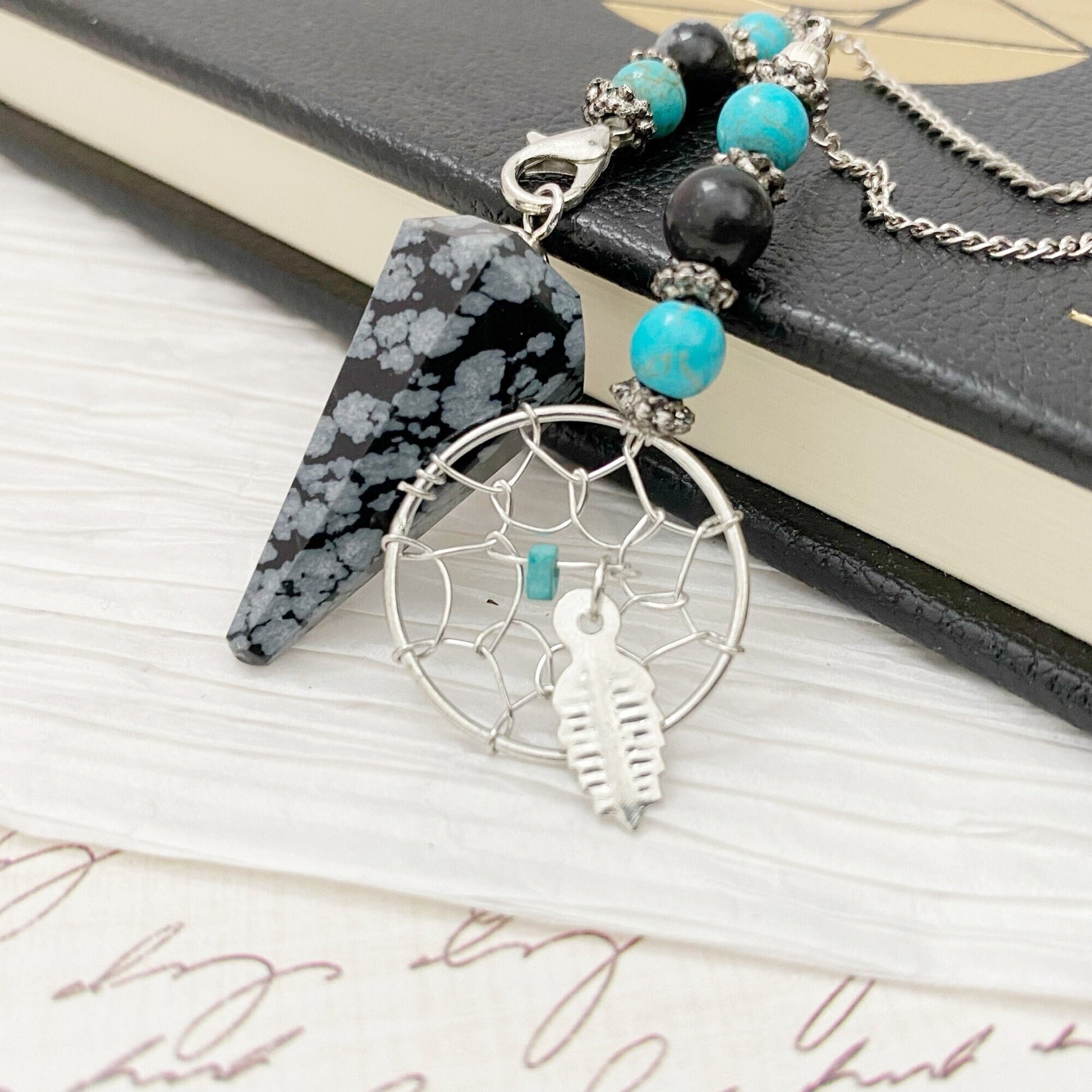 A necklace with a silver dreamcatcher charm pendant, featuring a small turquoise bead and a feather charm, lies on a black book with a geometric design. The necklace also includes a Snowflake Obsidian Hexagonal Pendulum and additional turquoise beads.