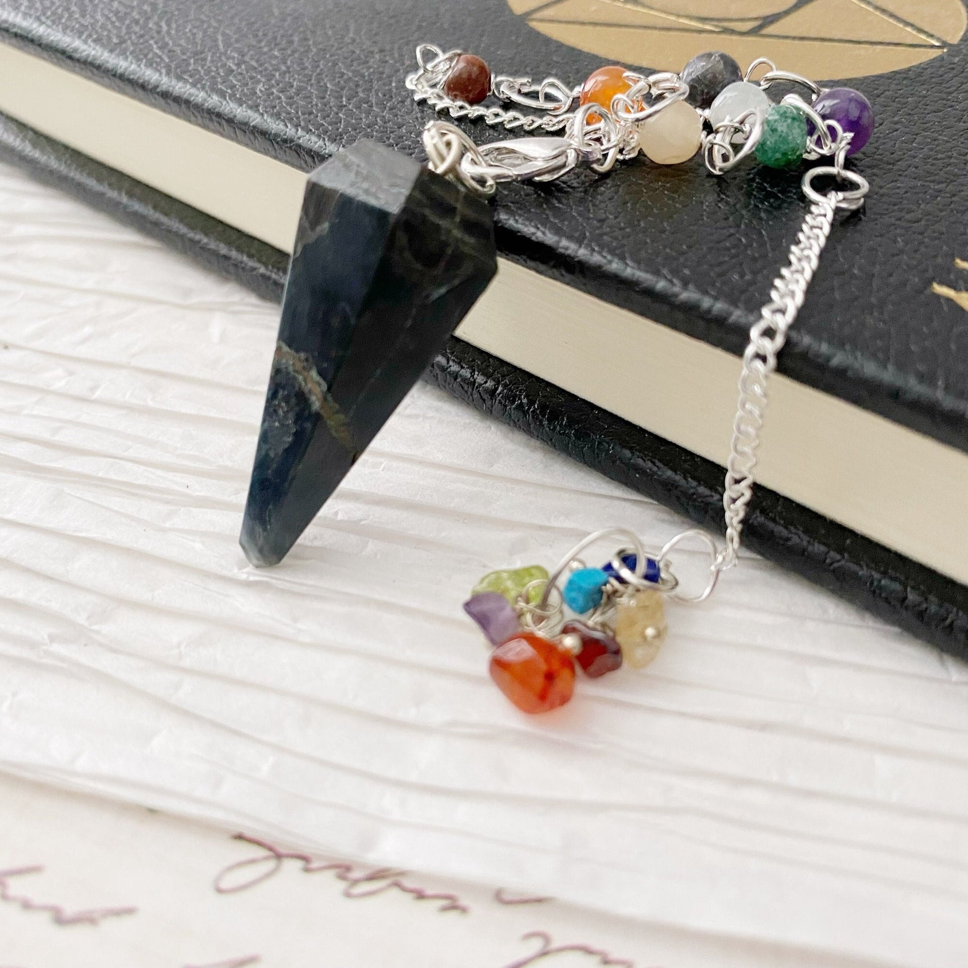 A photo of a Blue Tigers Eye Hexagonal Pendulum with a silver chain, adorned with small multicolored crystals, lying on a closed journal. The journal's cover has a subtle embossed pattern and is partially visible in the background. The setting includes white crinkled fabric.