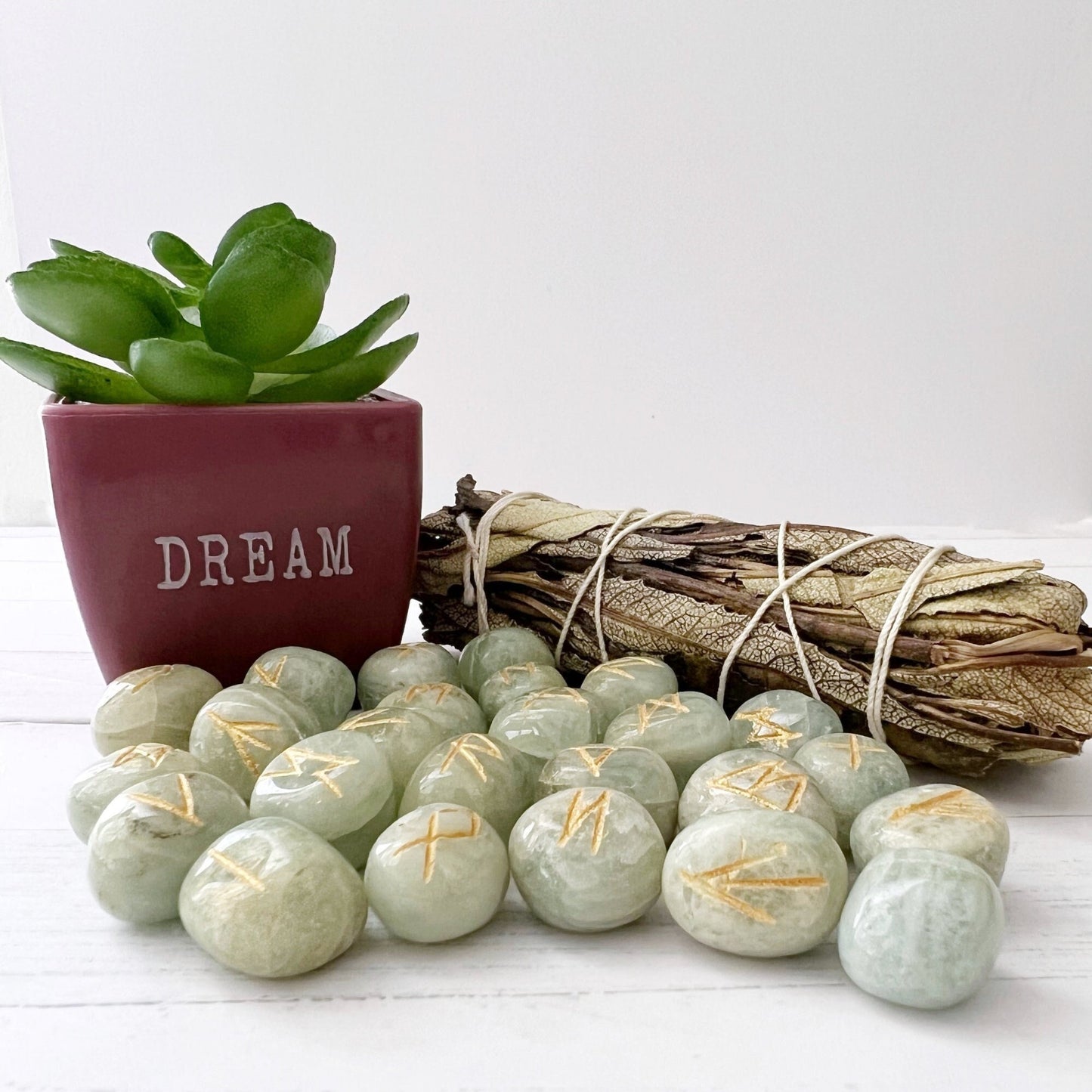 A maroon pot with a succulent plant and the word "DREAM" sits on a white surface. In front, there are pale green Aquamarine Elder Futhark Runes with gold markings. A bundle of sage tied with string is placed beside the stones.