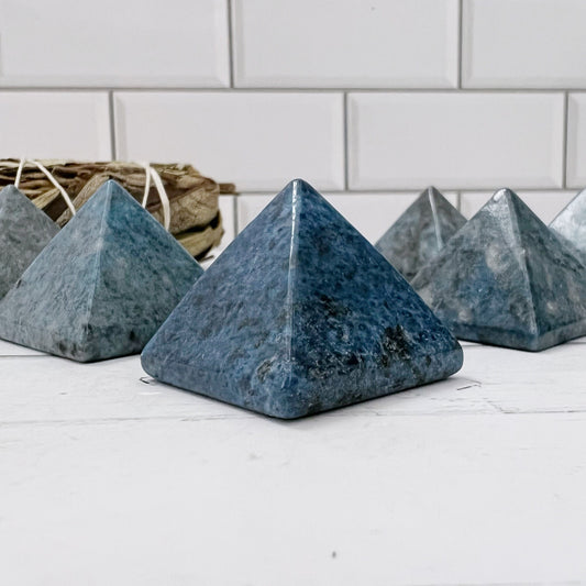 A collection of smooth, 1" Mini Dumortierite Pyramids, known for enhancing psychic abilities and Third Eye Chakra activation, are displayed on a white surface with a white tiled wall in the background. A basket of coiled rope is partially visible in the background. The focus is on a central pyramid in the foreground.