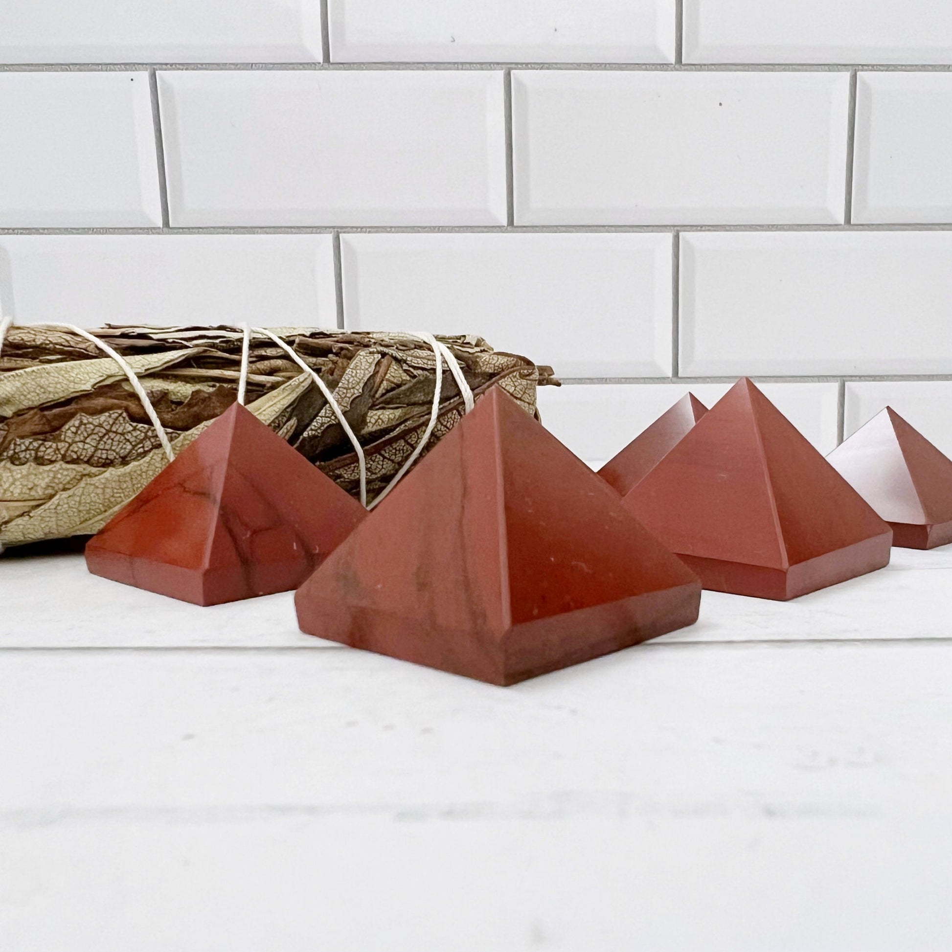 The image features several polished, reddish-brown 1" Mini Red Jasper Pyramids arranged on a white surface. In the background, there's a bundle of dried sage wrapped in string. The setting appears to be a minimalist space with a white tiled wall, ideal for harnessing kundalini energy and balancing the Base Chakra.