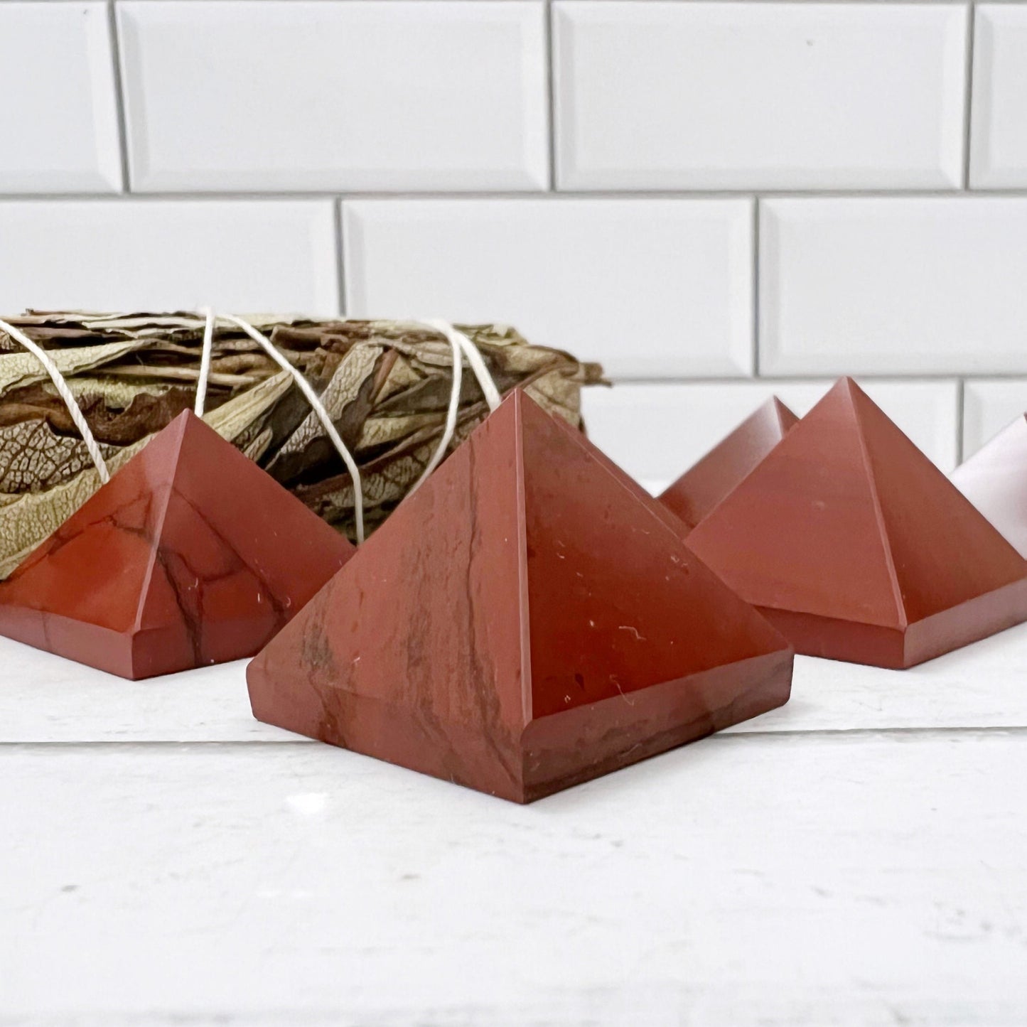 The image features several polished, reddish-brown 1" Mini Red Jasper Pyramids arranged on a white surface. In the background, there's a bundle of dried sage wrapped in string. The setting appears to be a minimalist space with a white tiled wall, ideal for harnessing kundalini energy and balancing the Base Chakra.