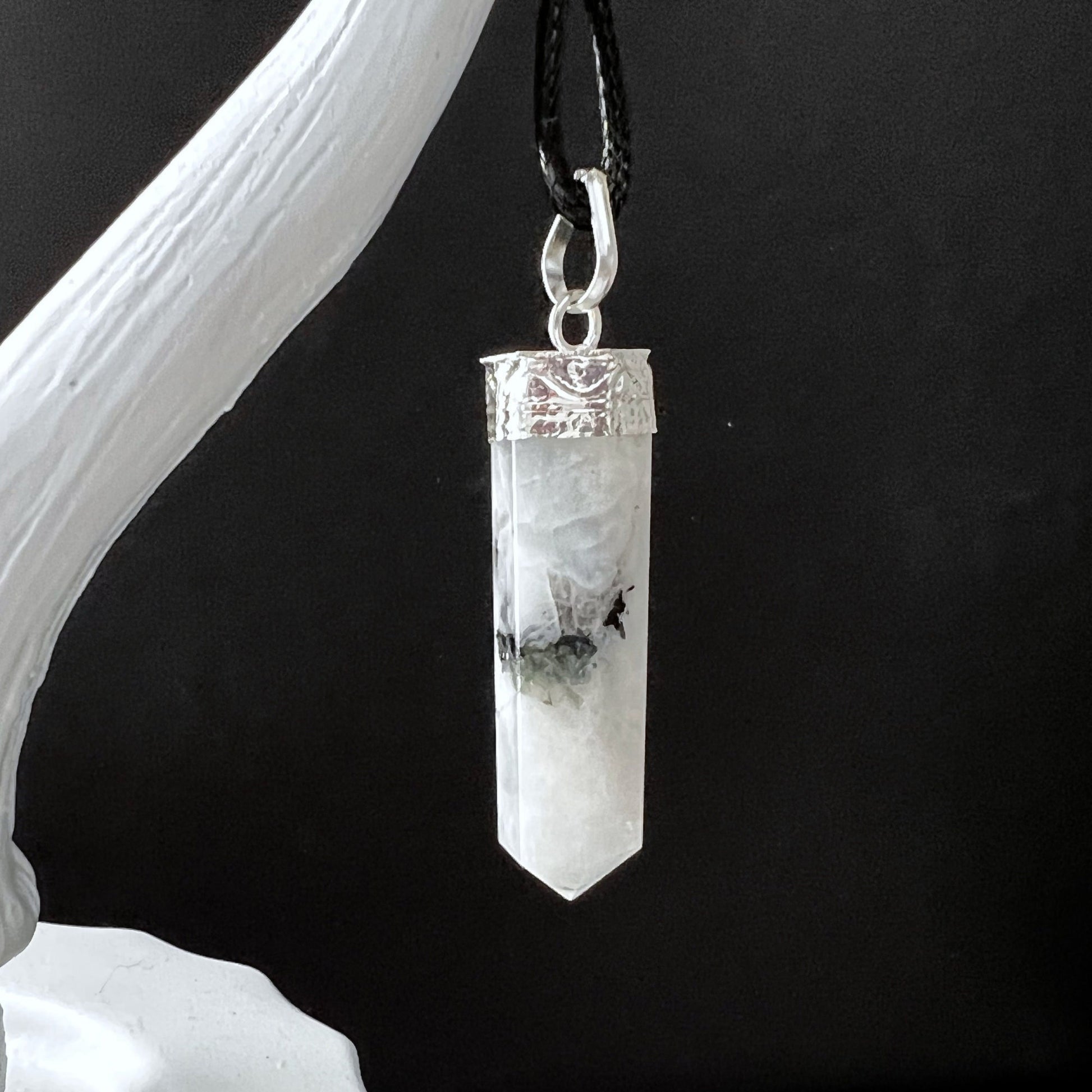 A hexagonal, pointed Rainbow Moonstone Point Pendant hangs from a black cord. The crystal is white with black inclusions and is capped with a silver metal top, featuring ornate detailing for psychic protection. It is suspended from a white, branch-like structure against a dark background.