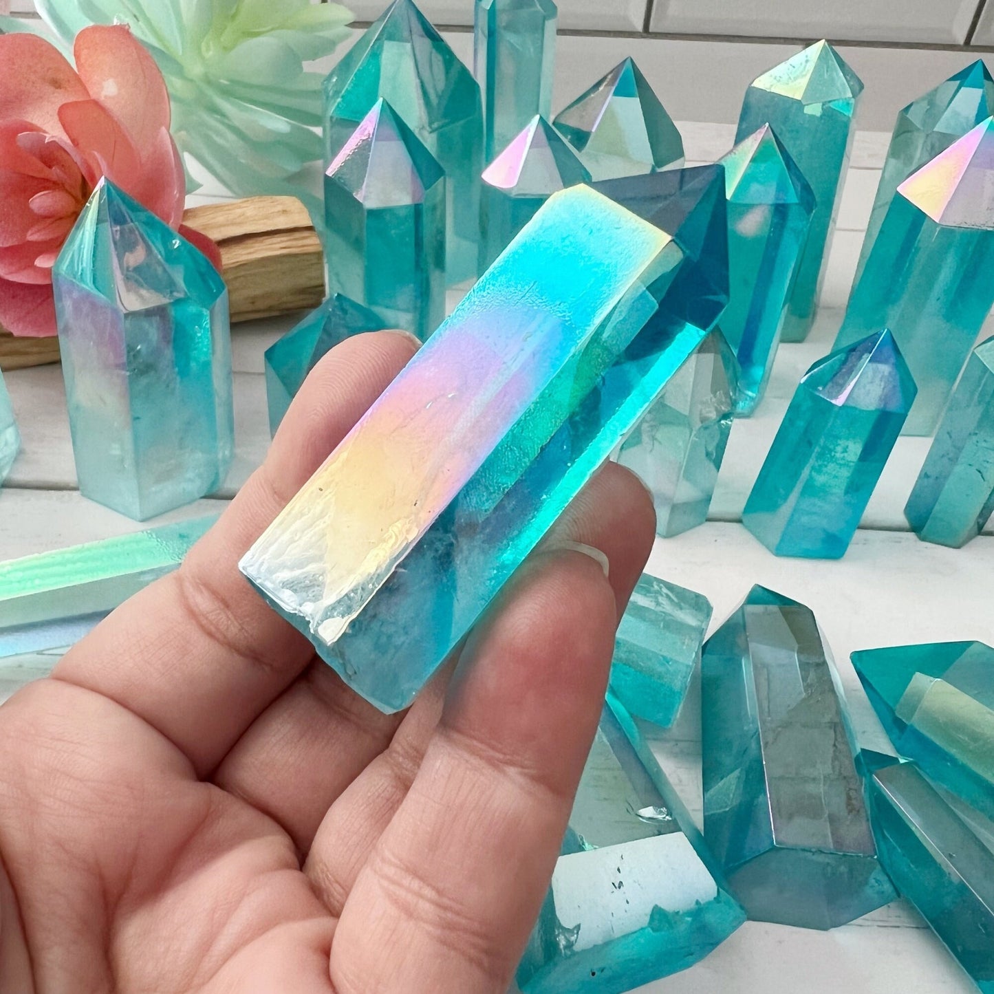 A hand holds a blueish-green, translucent 2-4" Polished Aqua Aura Quartz Point with a rainbow sheen. Numerous similar crystals of various sizes are displayed in the background on a white surface, alongside a decorative pink flower, forming an exquisite crystal grid reminiscent of an energy generator.