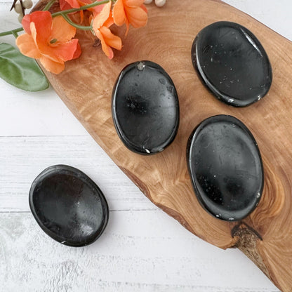 A wooden board adorned with three shiny Black Tourmaline Worry Stones and a cluster of orange flowers. A fourth worry stone rests on a white surface beside the board. The setup is bright and minimalistic, highlighting the smooth texture and protective qualities of the stones.