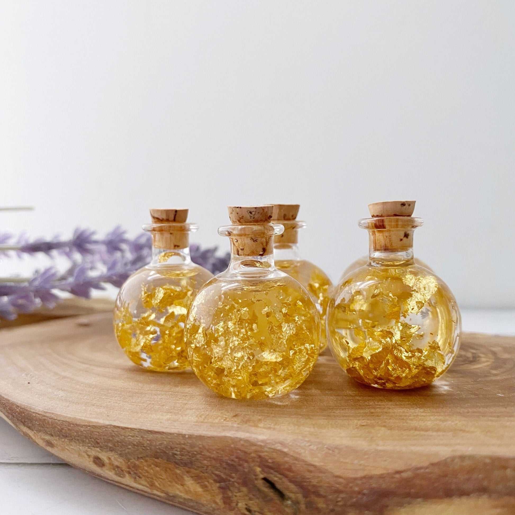 Four small, rounded 22K Gold Flakes Bottles with cork stoppers sit on a wooden surface. Each bottle contains 22K gold flakes and a transparent liquid. The background is minimalistic, with a hint of lavender flowers on the left side, perfect to attract wealth and elegance.