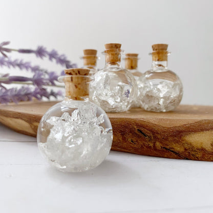 A .999 Silver Flakes Bottle filled with crystal-like white flakes and topped with a cork stopper, arranged on a wooden surface. In the background, there's a soft-focus sprig of purple lavender flowers.