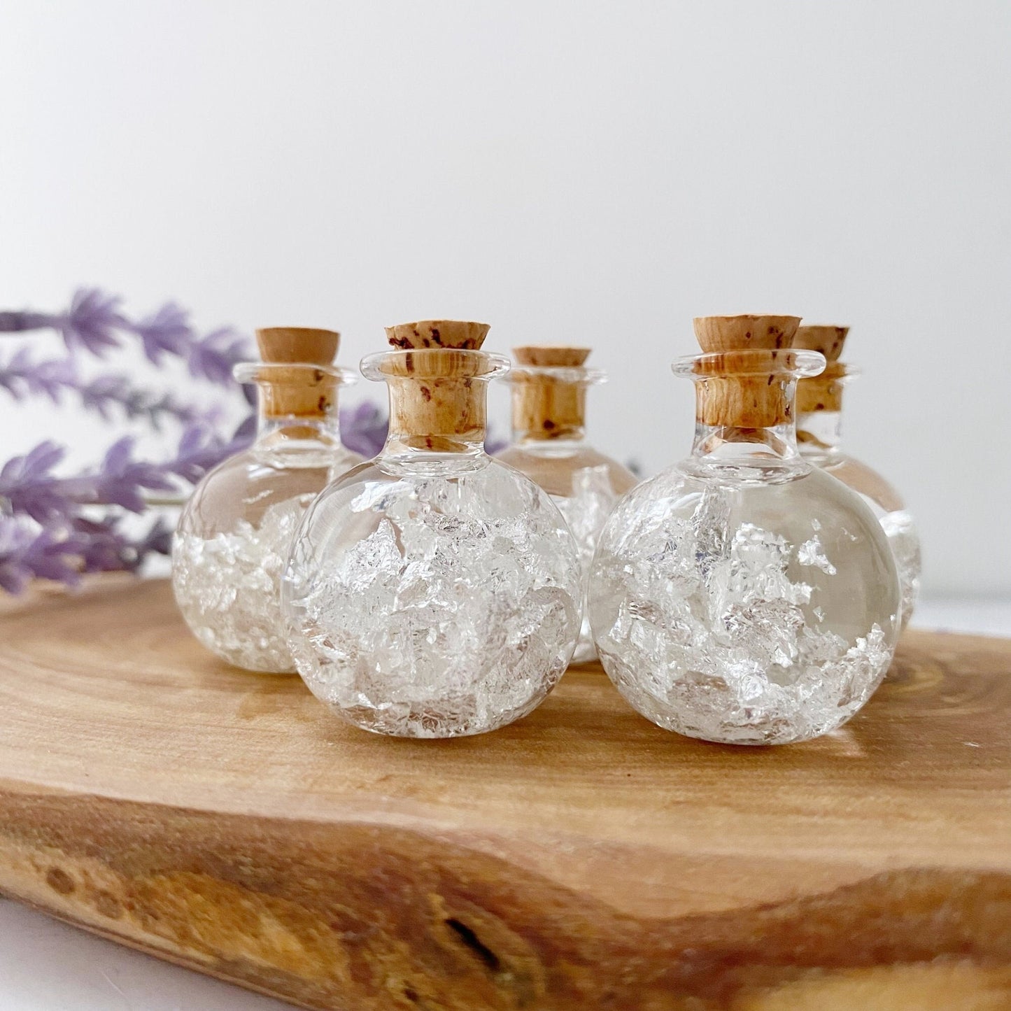 A .999 Silver Flakes Bottle filled with crystal-like white flakes and topped with a cork stopper, arranged on a wooden surface. In the background, there's a soft-focus sprig of purple lavender flowers.