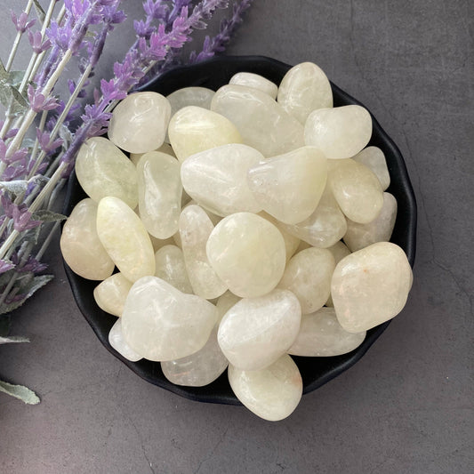 A black bowl filled with smooth, polished white stones and a piece of Natural Lemon Quartz Tumbled Stones sits on a dark gray surface. Sprigs of lavender flowers lie beside the bowl, evoking prosperity and abundance in the calm, natural ambiance.