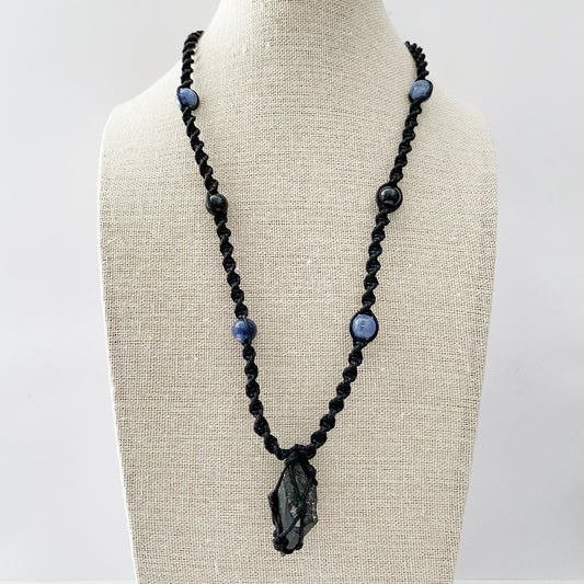 A handcrafted Black Tourmaline Crystal Point Net Cage Necklace displayed on a beige mannequin bust. The necklace features a black cord intricately knotted with spherical blue and black beads. At the center hangs a polished black crystal wrapped in black cord, showcasing skilled craftsmanship and protective energies.