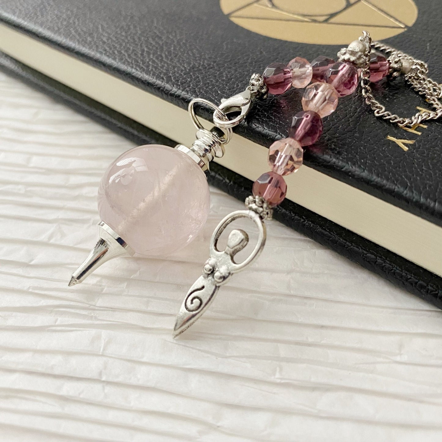 A Rose Quartz Crystal Ball Pendulum w/Goddess Charm with an intricate metal design sits on a black book adorned with a golden geometric pattern. The pendulum, featuring pink and purple beads along the chain, culminates in a small metal goddess charm.