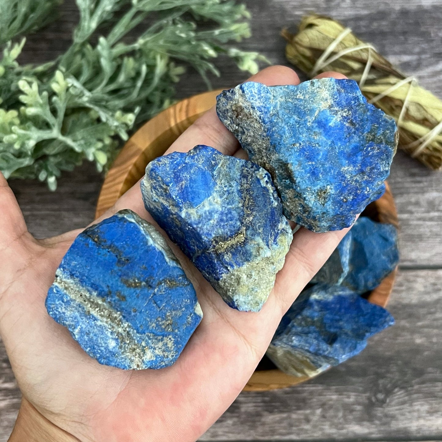 A wooden bowl filled with vibrant chunks of Raw Lapis Lazuli stones, known for their Throat Chakra activation, sits on a wooden surface. To the right of the bowl, a sage bundle wrapped in string lies serenely. In the background, a sprig of green foliage adds to the scene's tranquil ambience.