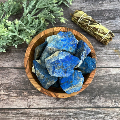 A wooden bowl filled with vibrant chunks of Raw Lapis Lazuli stones, known for their Throat Chakra activation, sits on a wooden surface. To the right of the bowl, a sage bundle wrapped in string lies serenely. In the background, a sprig of green foliage adds to the scene's tranquil ambience.