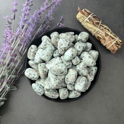 A bowl filled with polished Kiwi Jasper Tumbled Stones lies on a dark surface, their white tones flecked with black specks. To the left, sprigs of purple lavender rest beside the bowl. In the upper right corner, a bundle of dried herbs tied with string completes this serene scene designed for stress relief and warding off negative energy.