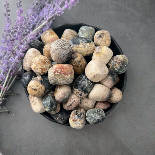 A small black bowl filled with smooth, polished stones of various earthy colors, including Fossilized Palm Root Tumbled Stones in tan, black, and gray, sits on a gray surface. Delicate lavender sprigs lie beside the bowl, adding a touch of purple to the tranquil scene.