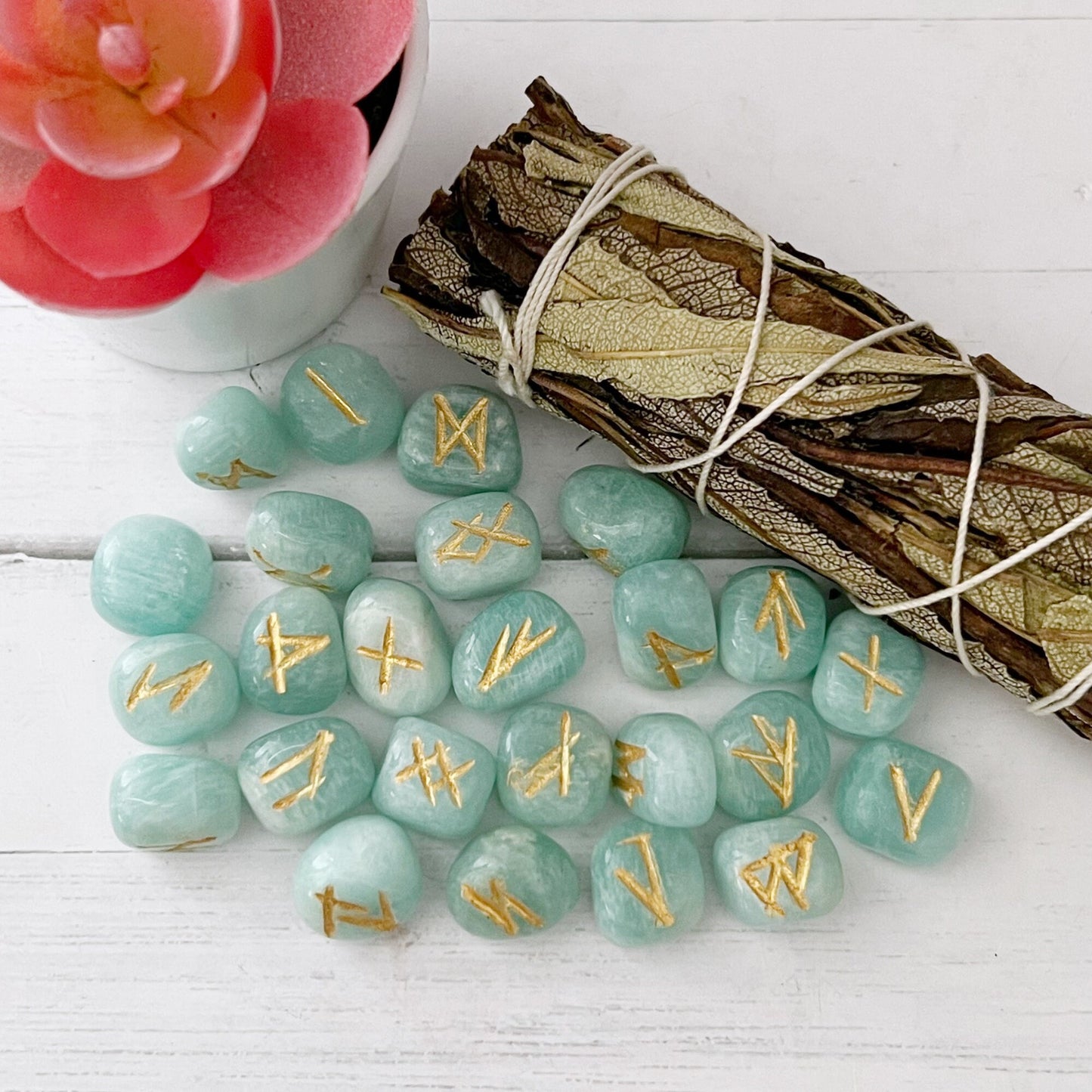 A potted pink succulent next to a bundle of dried sage and a collection of hand-carved Amazonite Elder Futhark Runes are placed on a white wooden surface. The stones are scattered in the foreground.