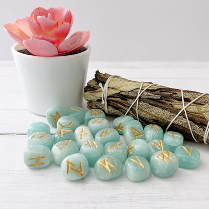 A potted pink succulent next to a bundle of dried sage and a collection of hand-carved Amazonite Elder Futhark Runes are placed on a white wooden surface. The stones are scattered in the foreground.