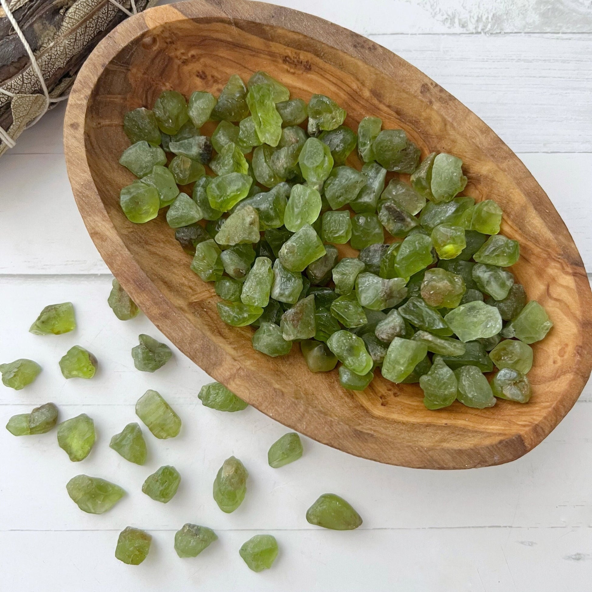 A wooden bowl filled with numerous small, roughly shaped green stones, likely natural peridot stones or crystals, placed on a light-colored surface. Several Raw Peridot Crystal (1 GM Mini) are scattered beside the bowl. The bowl has a rustic, natural appearance.