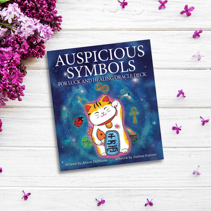 The "Auspicious Symbols for Luck and Healing Oracle" book rests on a white wooden surface adorned with lilac blooms and scattered purple flowers. The cover features a colorful illustration of a beckoning cat and various symbols against a blue background, radiating healing energy.