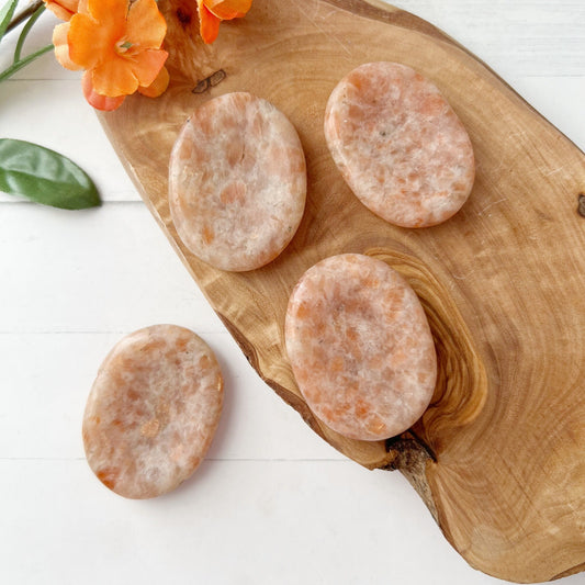 Four oval-shaped polished Sunstone Worry Stones, perfect for enhancing intuition, are displayed on a wooden board with a natural grain pattern. An orange flower with green leaves is positioned in the upper left corner, adding a touch of color to the composition. The background is a white surface.