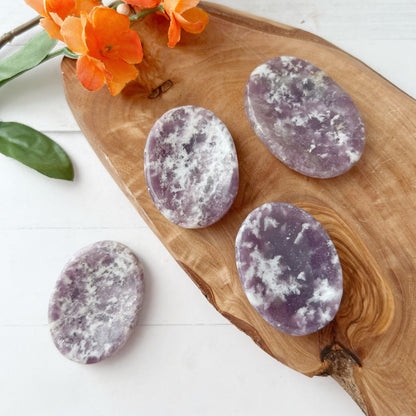 A wooden board with four polished Lepidolite Worry Stones displaying white crystal patterns is shown. An orange blossom branch is placed at the top of the board, adding a touch of color to the serene arrangement. The background is a white surface, enhancing the sense of emotional healing and cosmic awareness.